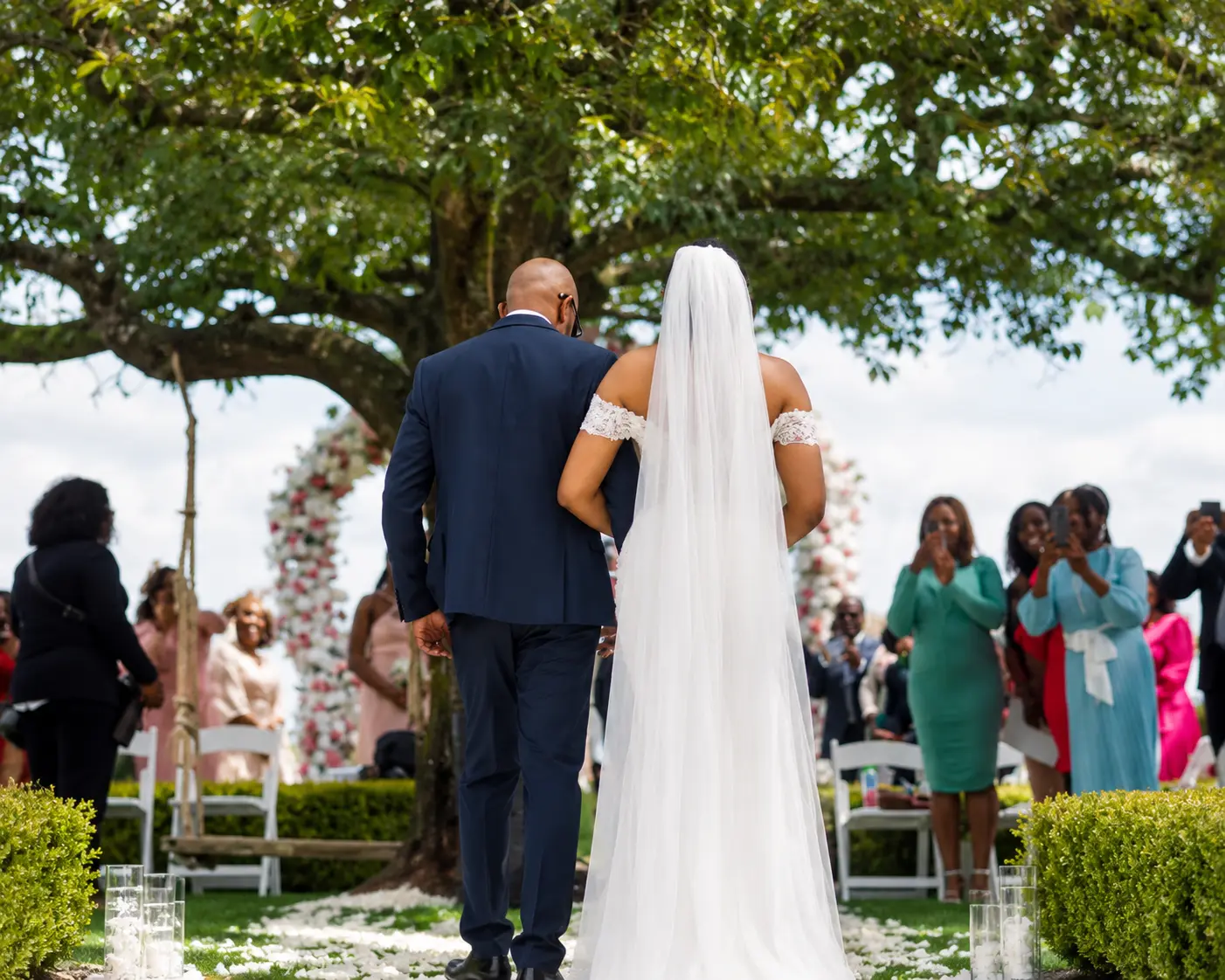 Bride walking down a garden aisle on her father's arm beneath a tree